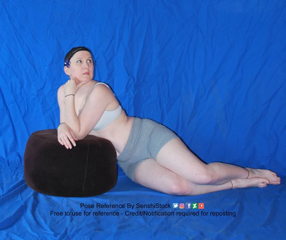 A woman in grey athletic wear lies on a blue fabric backdrop, propped up on a large brown beanbag, looking upward with her…