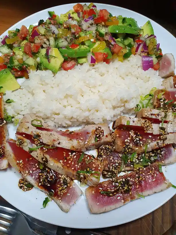 A white plate holds a colorful poke bowl with sliced seared tuna topped with sesame seeds and green onions, a mound of white…