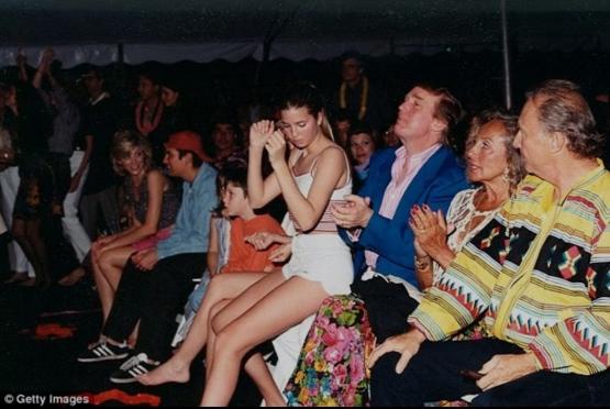 A candid photo of a crowd at an event, featuring a woman in a white dress with her hands clasped near her face, appearing…