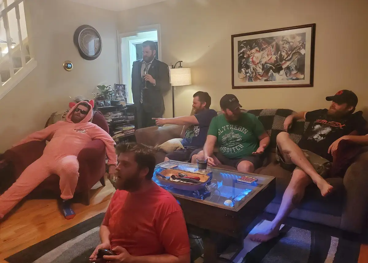 A group of men are gathered in a living room, one in a pink pig onesie lounging in a chair, another in a suit standing in…