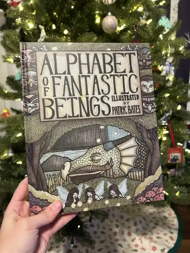 A hand holds up a children's book titled 'Alphabet of Fantastic Beings' by Patric Bates, featuring a dragon sleeping under…