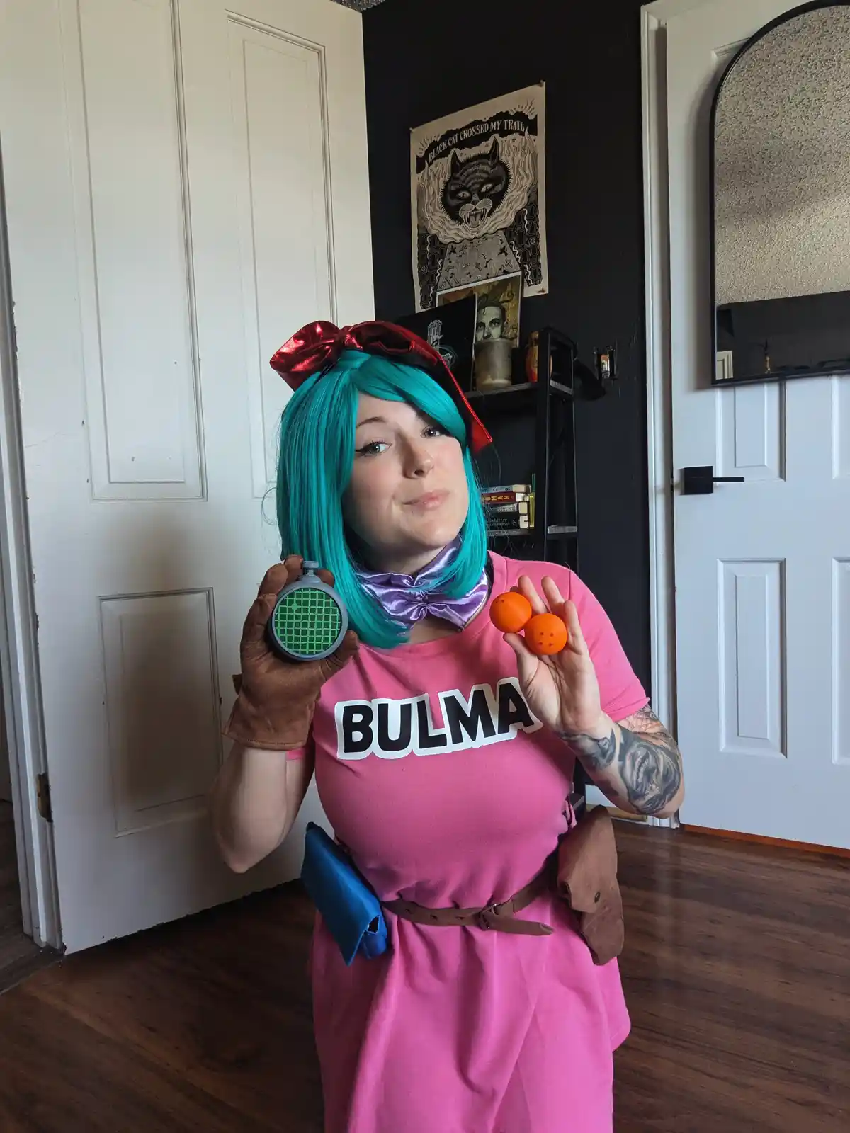 A person in a Bulma cosplay from Dragon Ball Z, wearing a pink dress with 'BULMA' printed on it, a purple bow tie, and a red…