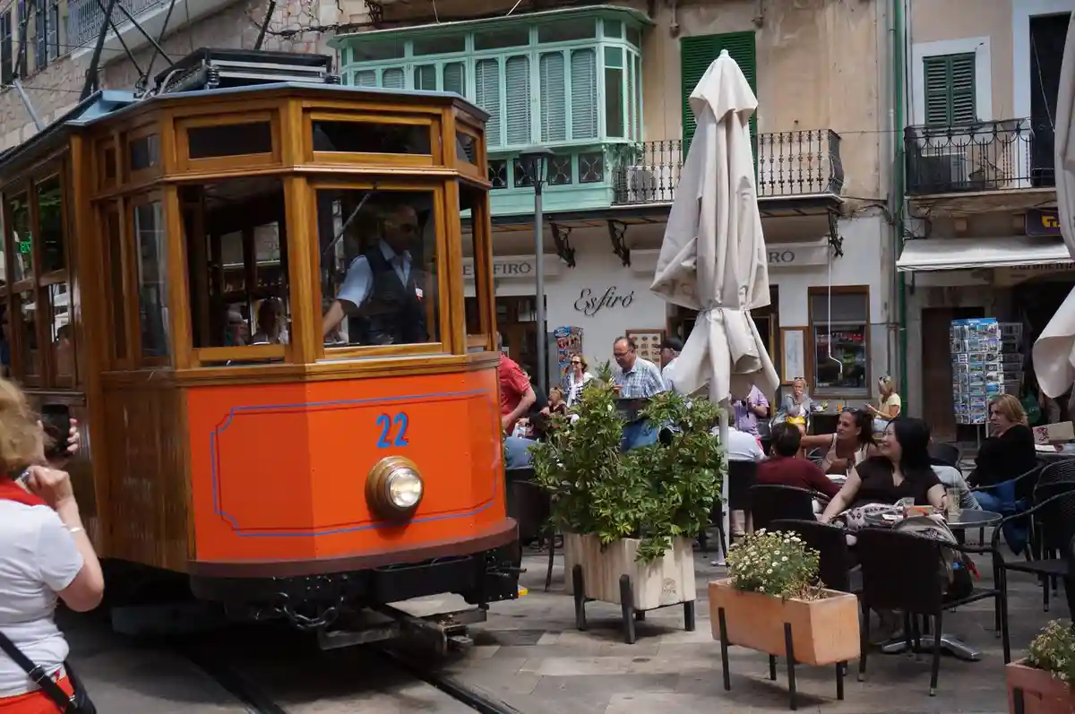 An orange and wooden vintage tram, numbered 22, moves through a narrow European street lined with cafes where people are…