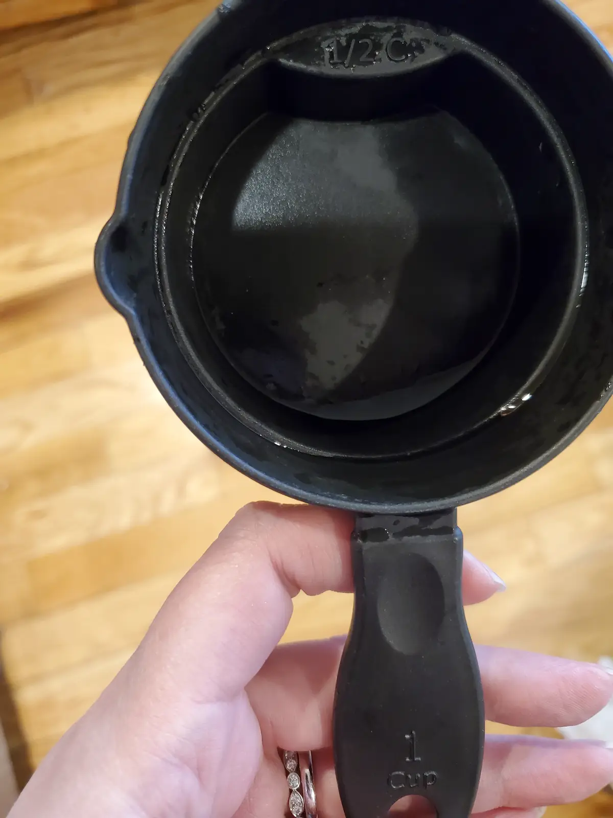 A hand holds a black plastic 1 cup measuring cup, showing its interior which has a 1/2 C marking. The cup is visibly dirty…