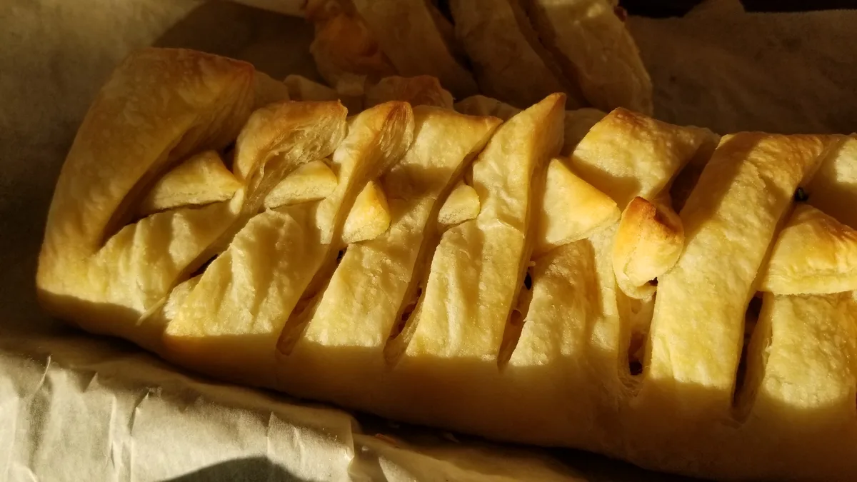 A close-up photo of a golden-brown, flaky pastry with a lattice top, resting on parchment paper. The overlaid text reads…