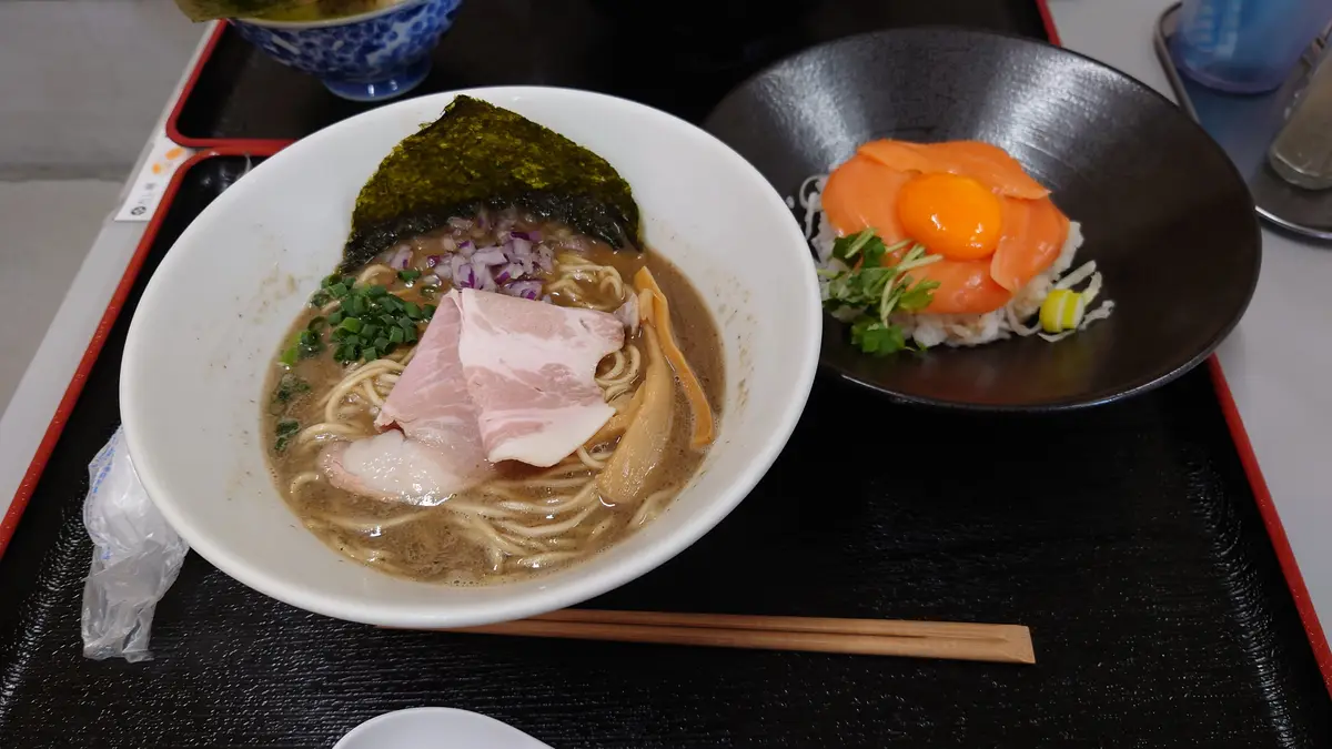 A close-up photo shows a white bowl of ramen with broth, noodles, pork, green onions, red onions, and nori seaweed, next to…