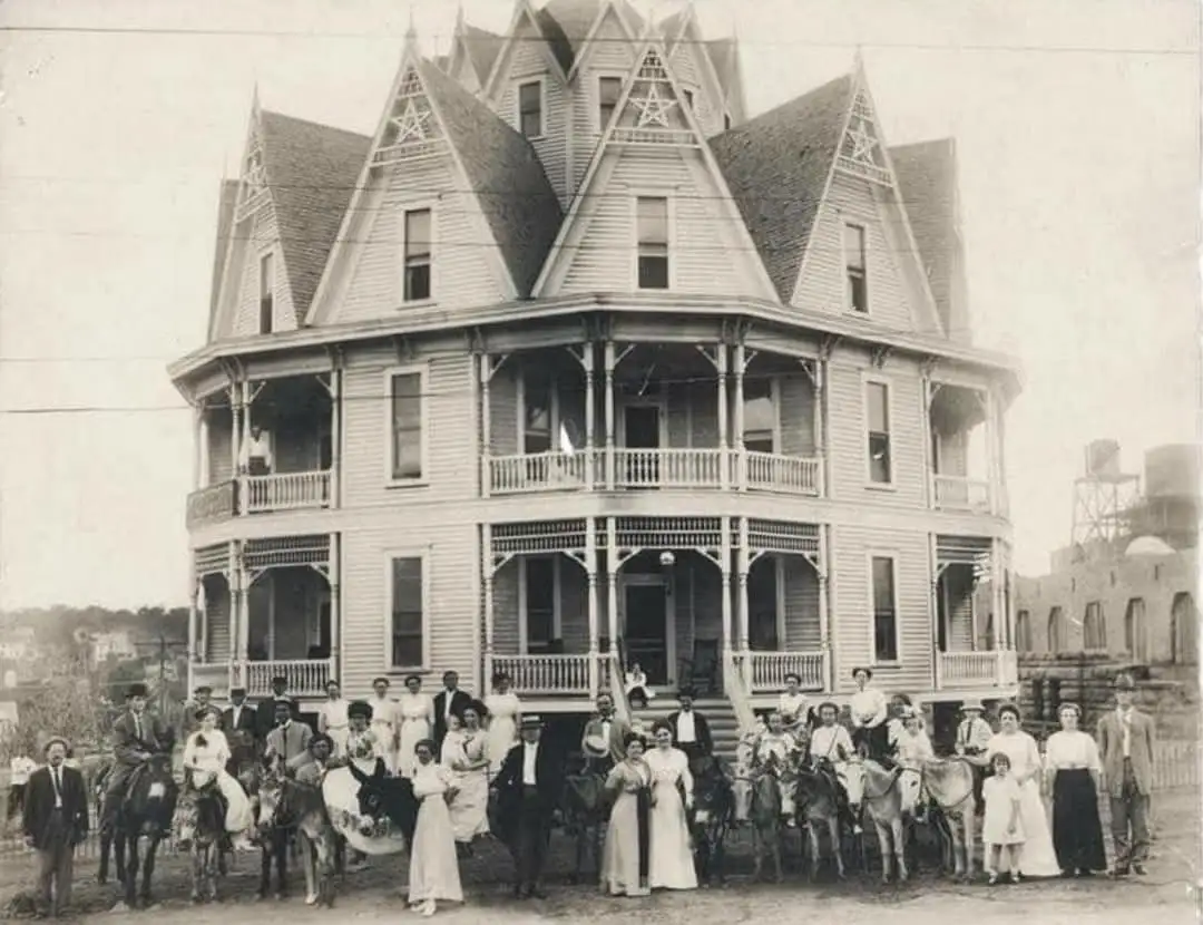 A sepia-toned historical photograph shows a large, ornate Victorian house with multiple gables and balconies, with a group…