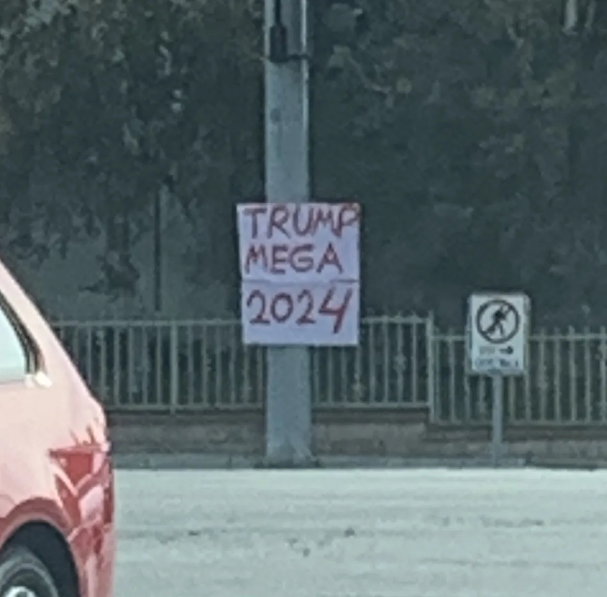 A handmade sign taped to a utility pole reads 'TRUMP MEGA 2024' in red marker. A red car is partially visible on the left…