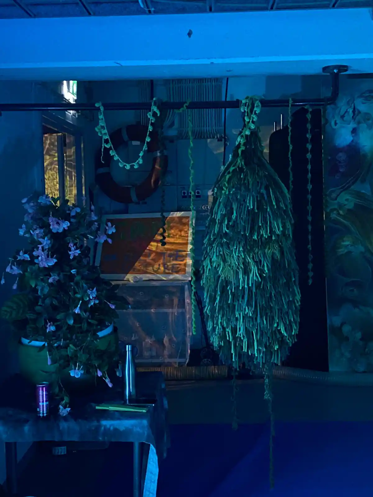 A dimly lit room bathed in blue light features a large, green, fringed decorative hanging object resembling a mop head. To…