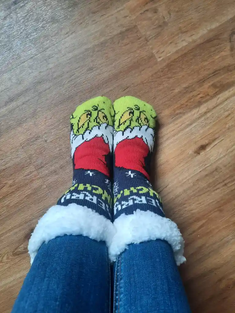 A person wearing festive, Grinch-themed socks with a green Grinch face, red Santa hat, and white fluffy trim, sitting on a…