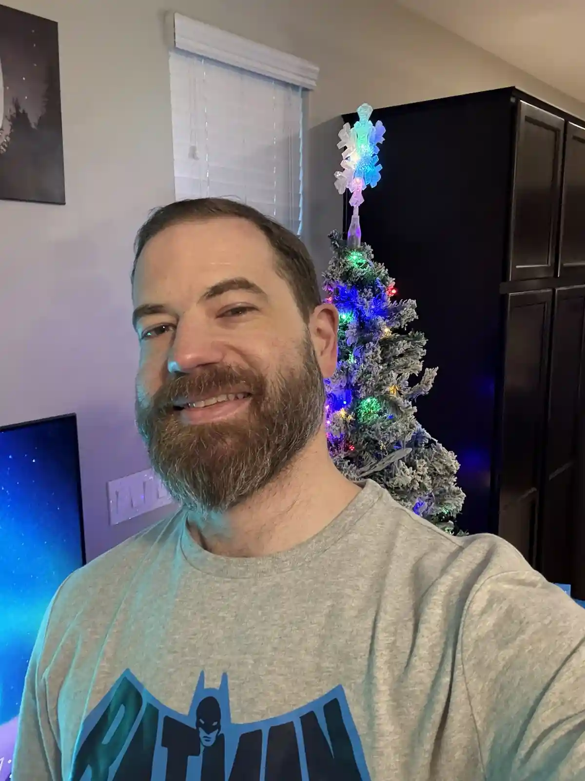 A man with a beard and Batman t-shirt smiles at the camera while standing in front of a small, decorated Christmas tree with…