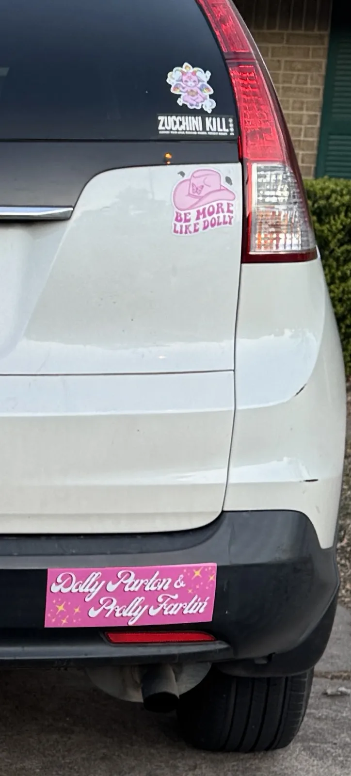 The rear of a light-colored SUV displays three stickers: a 'ZUCCHINI KILL' sticker near the top, a pink cowboy hat sticker…