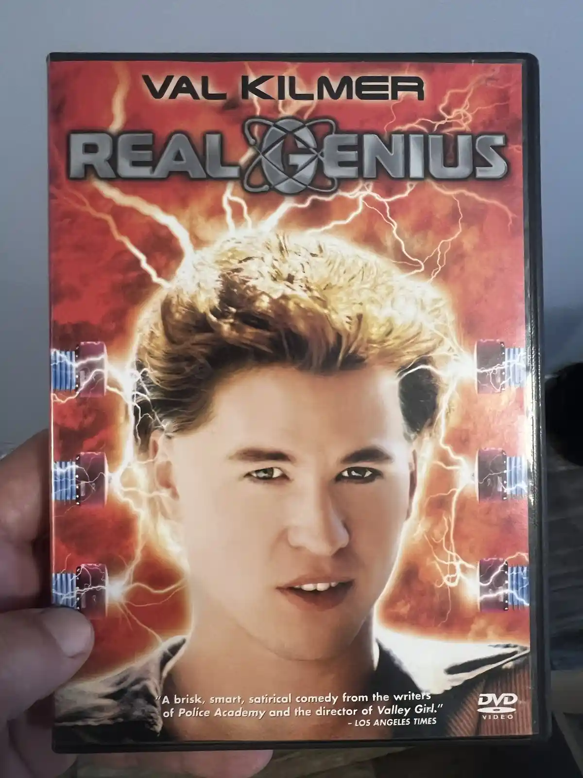 A hand holds a DVD cover for the 1985 film 'Real Genius' starring Val Kilmer, featuring his face with lightning bolts around…