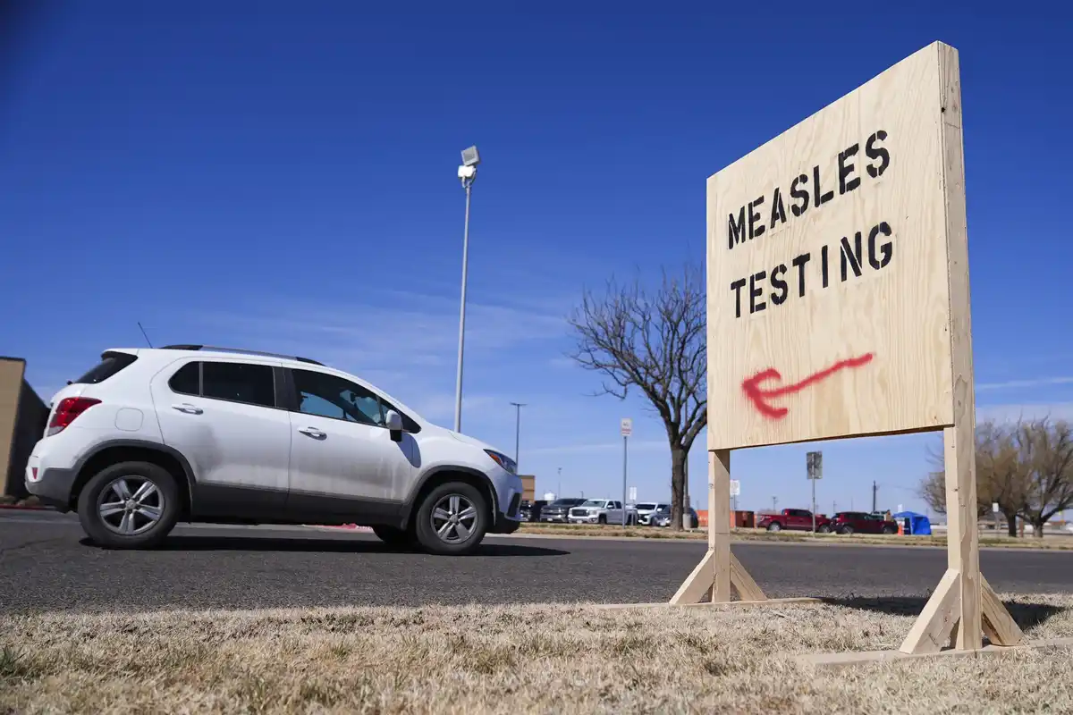 A white SUV drives past a wooden sign that reads 'MEASLES TESTING' with a red spray-painted arrow pointing left. The scene…