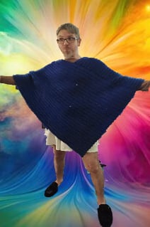 A person with glasses and a neutral expression stands with arms outstretched, wearing a large, dark blue knitted poncho or…