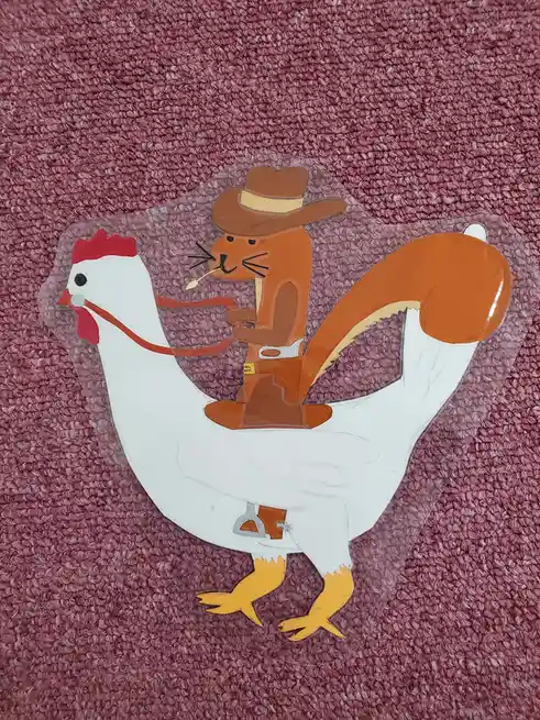 A clear plastic decal depicts a cartoon squirrel wearing a cowboy hat and smoking a pipe, riding a white chicken with red…