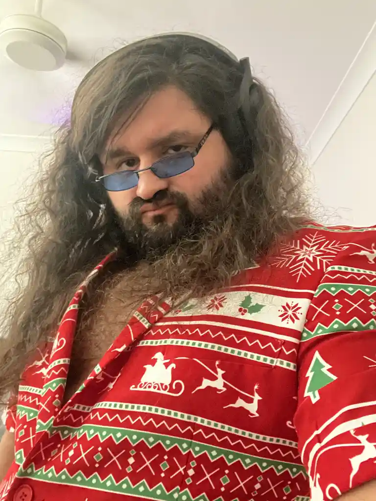 A man with long curly hair, a beard, and blue-tinted glasses wears a red Christmas-themed shirt with snowflakes, reindeer…