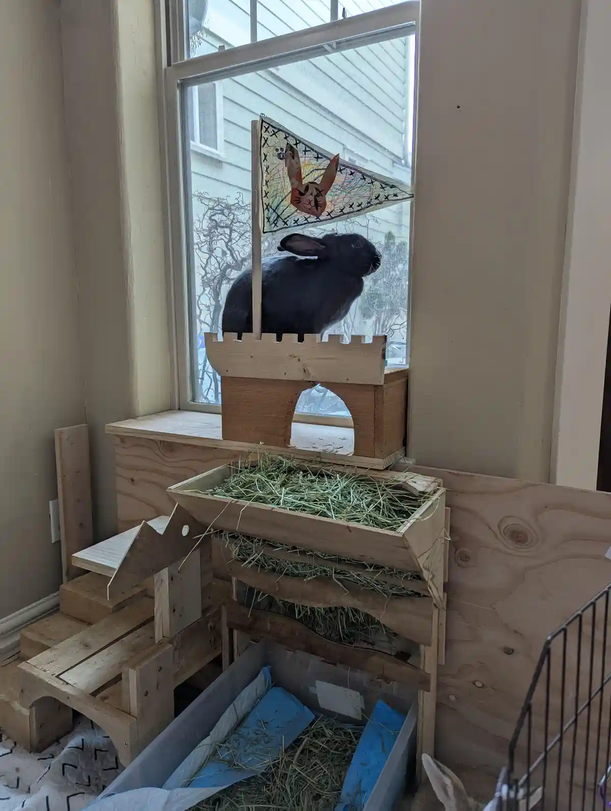 A black rabbit sits atop a wooden castle structure placed on a windowsill, looking out the window. A handmade flag with a…
