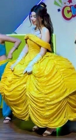 A person in a full yellow Belle costume from Beauty and the Beast, complete with gloves and a large gown, sits on a green…