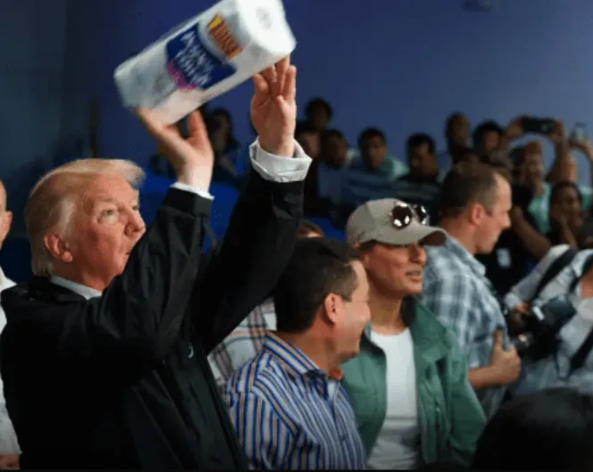 A man in a dark suit holds up a roll of Bounty toilet paper in a crowd, looking upward with an intense expression. The…