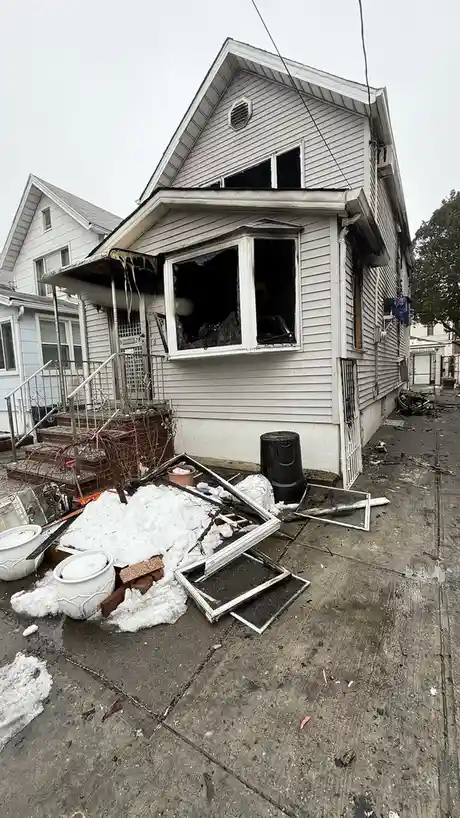 A damaged white house with broken windows and charred siding, surrounded by debris including shattered picture frames, snow…