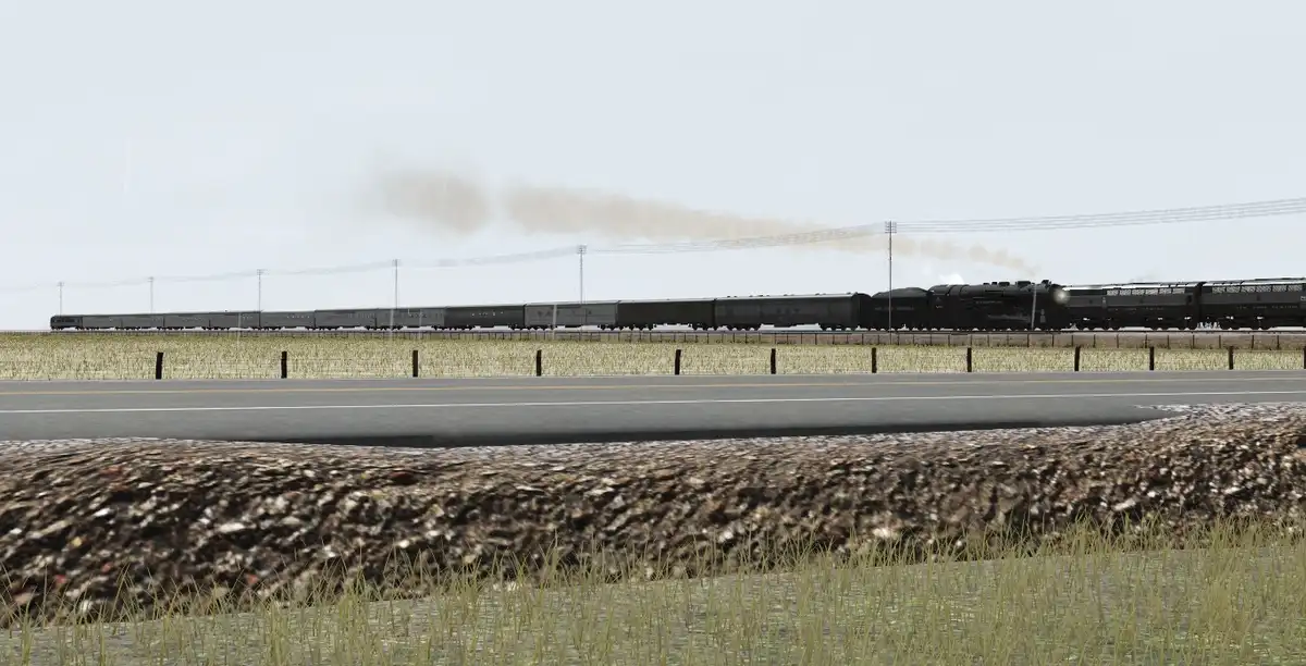 A long steam locomotive train emits smoke as it travels across a rural landscape under a gray sky, with a road and gravel…