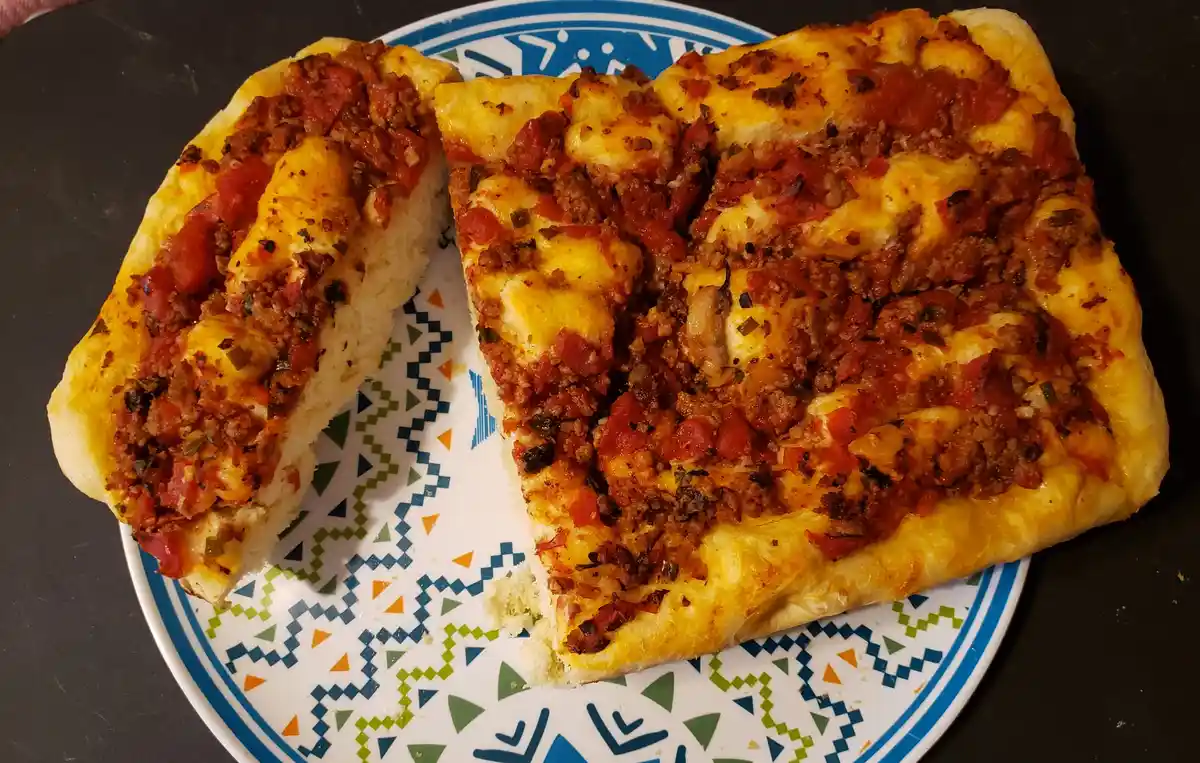 A close-up photo shows a thick, rectangular, bread-like item topped with melted cheese, tomato sauce, and ground meat…