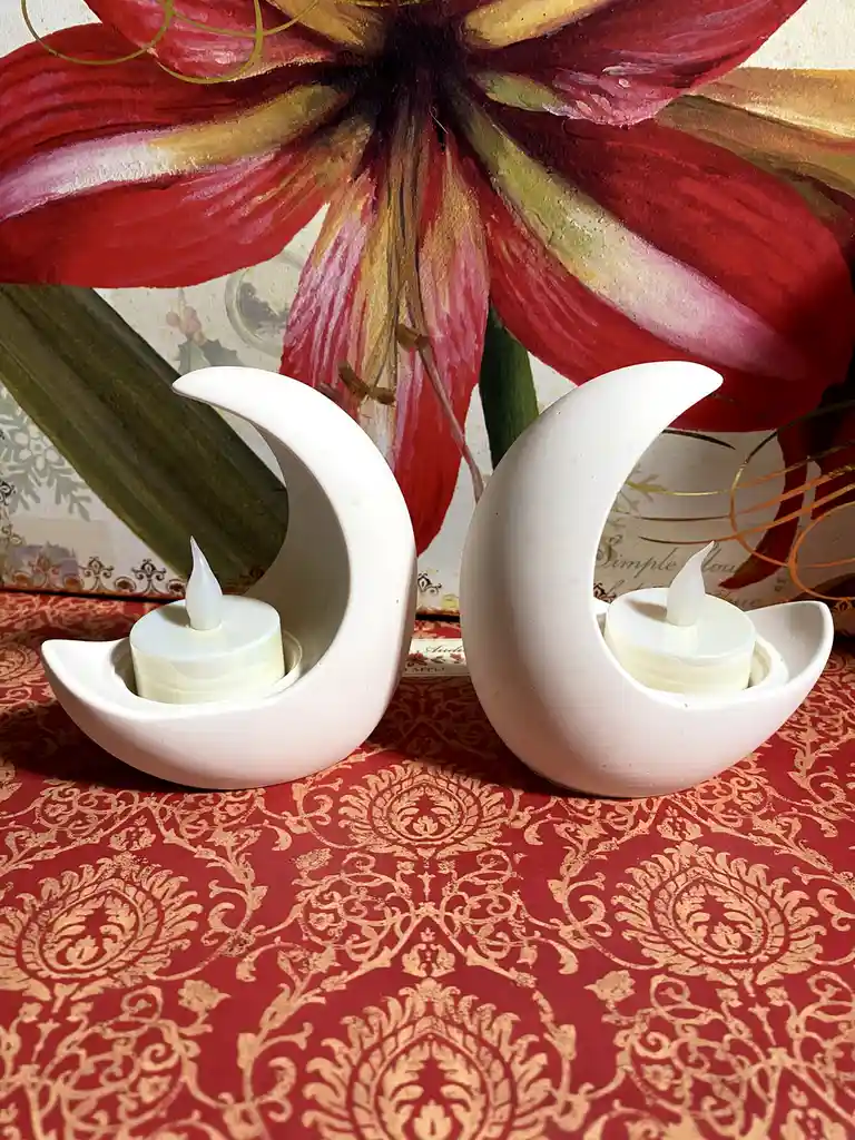 Two white ceramic moon-shaped candle holders, each containing a lit LED tea light, sit on a red and gold damask-patterned…