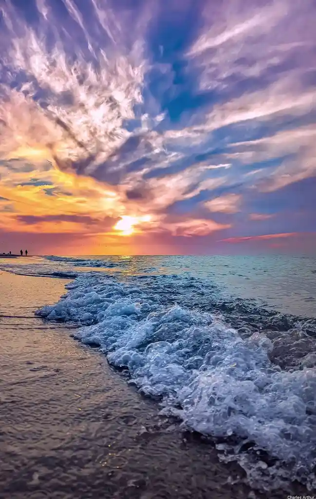 A serene beach sunset photo with vibrant orange, pink, and purple clouds above a calm ocean where gentle waves crash onto…
