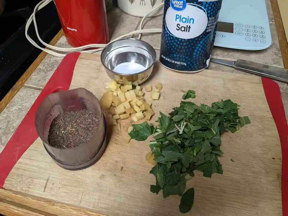 A kitchen counter shows a wooden cutting board with chopped ginger, a pile of fresh mint leaves, a small metal bowl of salt…