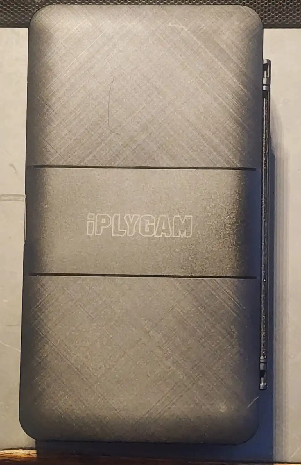 A close-up photo shows a dark gray, textured, rectangular device with a visible hinge on the right side, resembling a laptop…