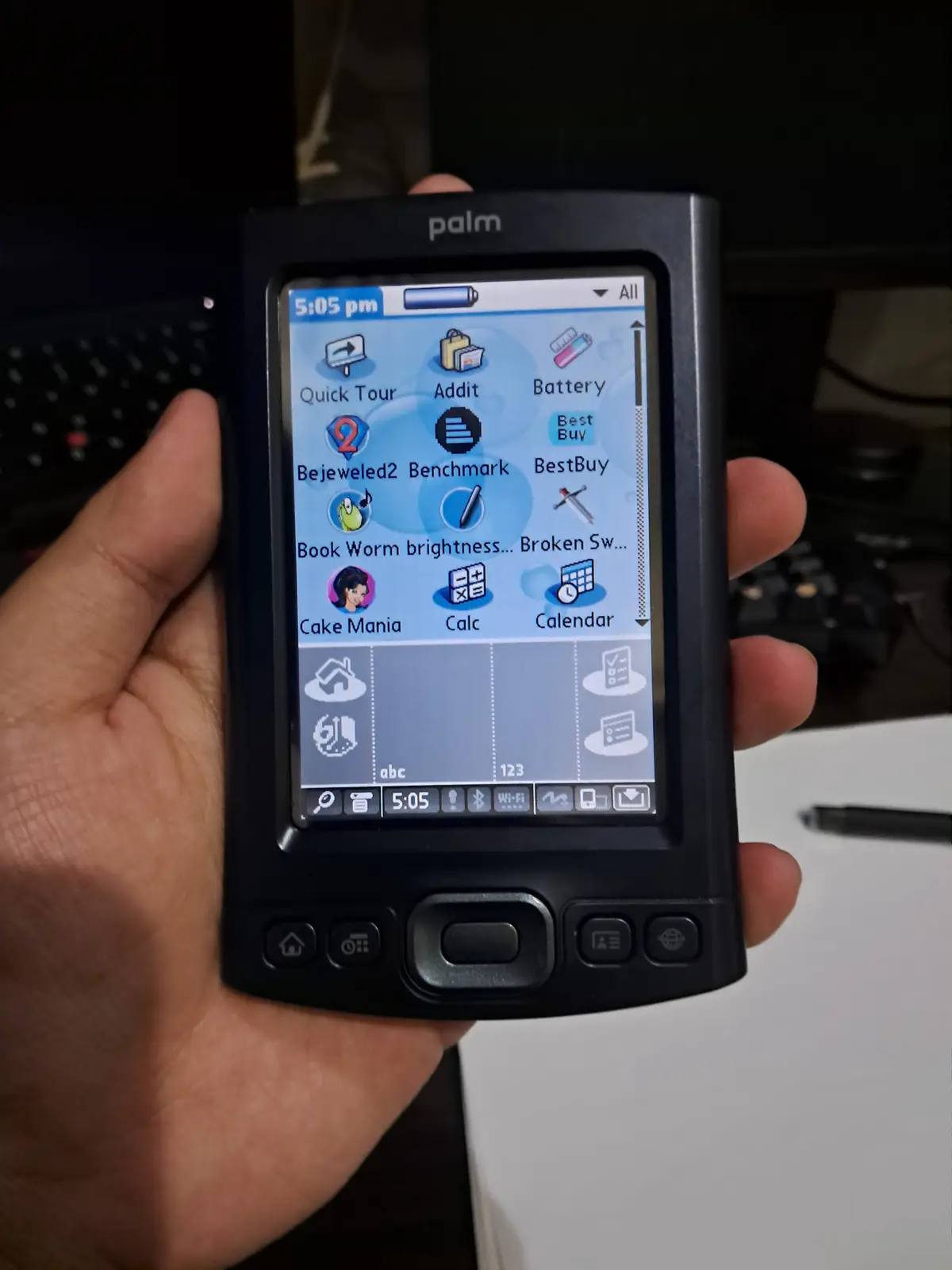 A hand holds a black Palm handheld device displaying its home screen with app icons like Bejeweled2, Cake Mania, and…