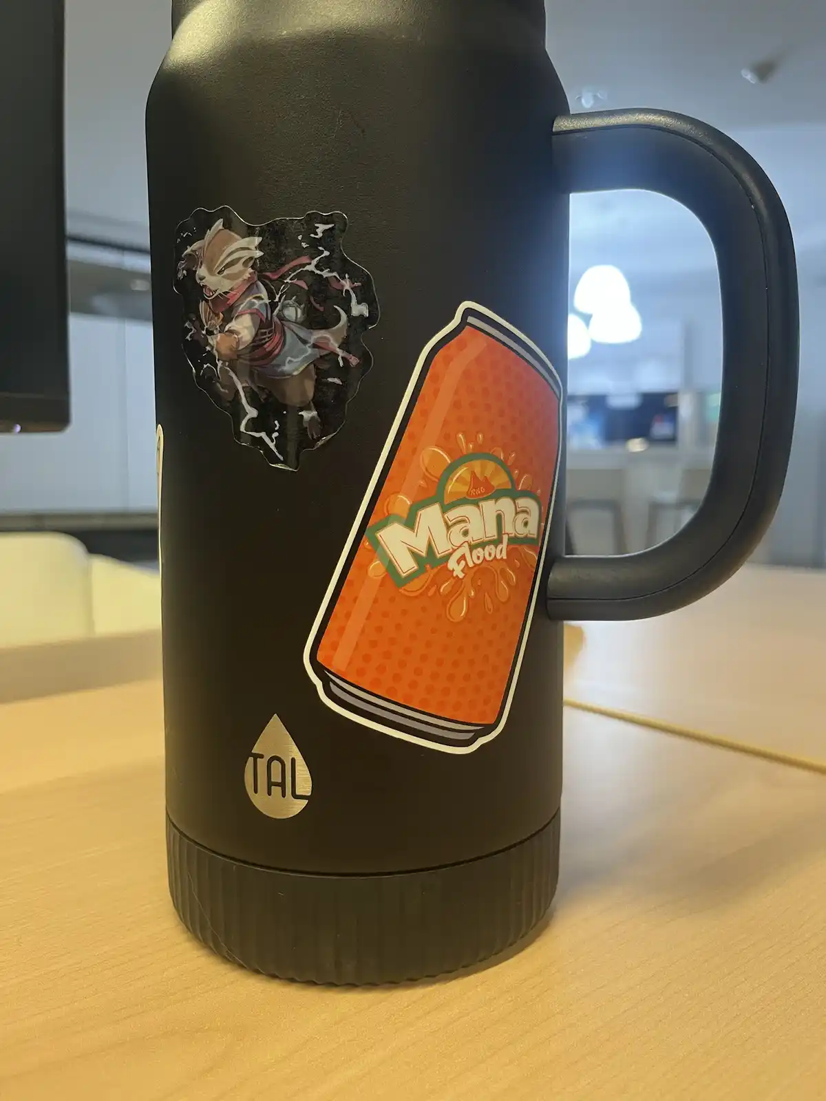 A black insulated mug with a handle sits on a wooden table, featuring two stickers: one of a cartoon fox-like character and…