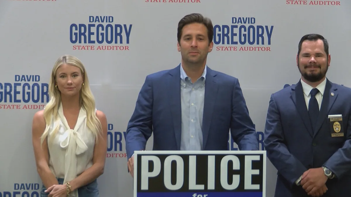Three people stand before a backdrop with 'DAVID GREGORY STATE AUDITOR' repeated. The central man stands behind a podium…
