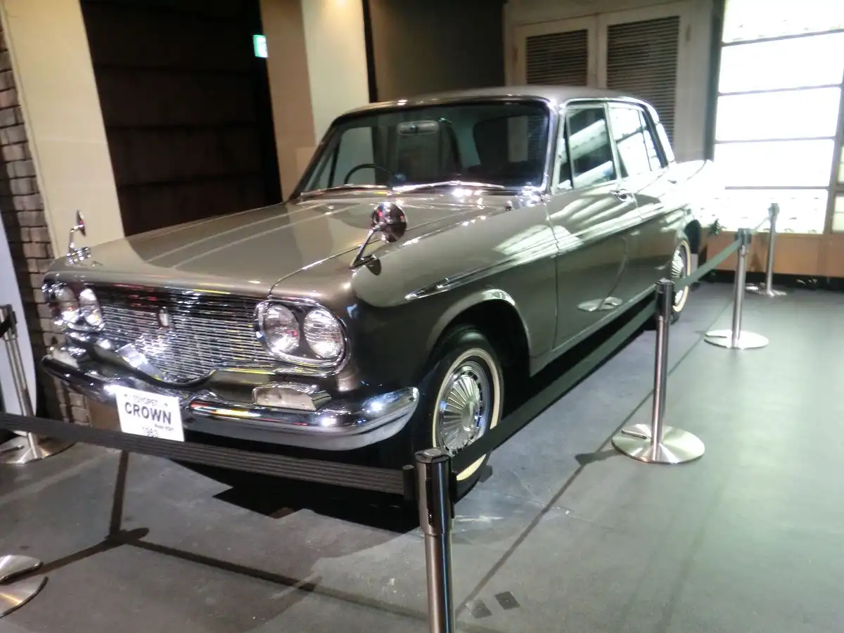 A vintage 1963 Toyota Crown sedan is displayed in a museum or showroom, surrounded by stanchions and ropes to keep viewers…