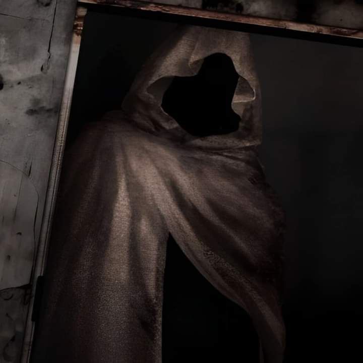 A dark, grainy image shows a shadowy figure in a tattered, hooded cloak standing in a doorway, with only the outline of…