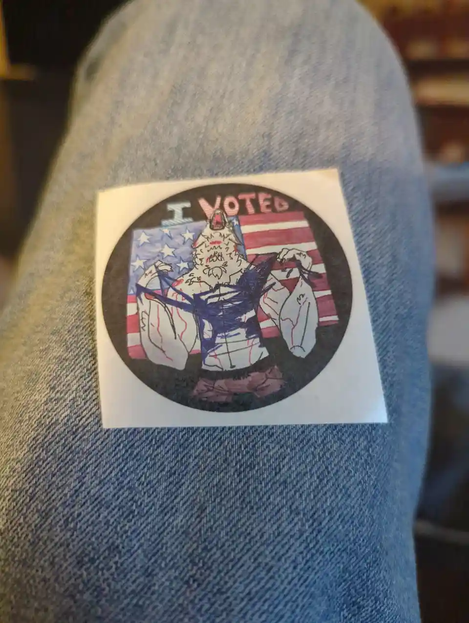 A hand-drawn, circular 'I VOTED' sticker is stuck on a person's denim jeans. The sticker features a cartoonish, muscular…