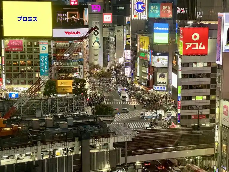 a high-angle nighttime photo of a crowded, brightly lit urban intersection in japan, likely shibuya, filled with towering…