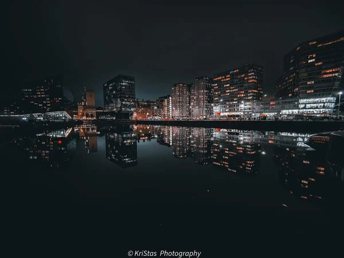 A nighttime cityscape photograph shows modern high-rise buildings with illuminated windows reflected in calm water below…