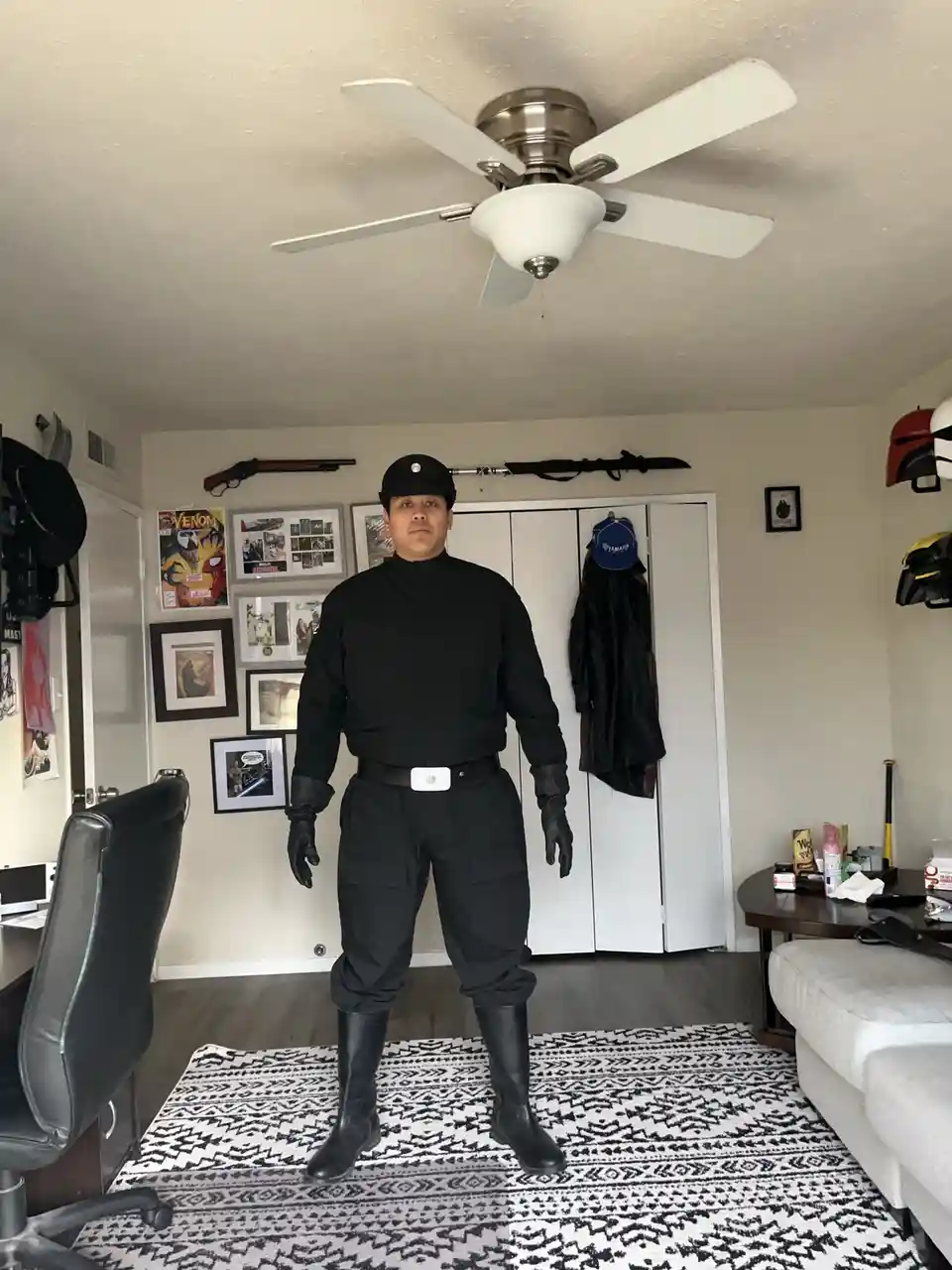 A man in a full black Stormtrooper costume from Star Wars stands in a bedroom, looking directly at the camera. The room has…