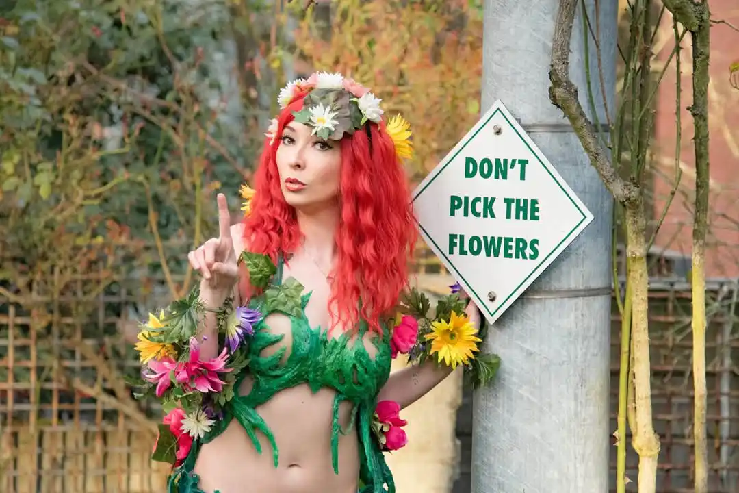 A woman dressed as Poison Ivy, with red hair, a floral crown, and a green leafy outfit, stands next to a sign that reads…