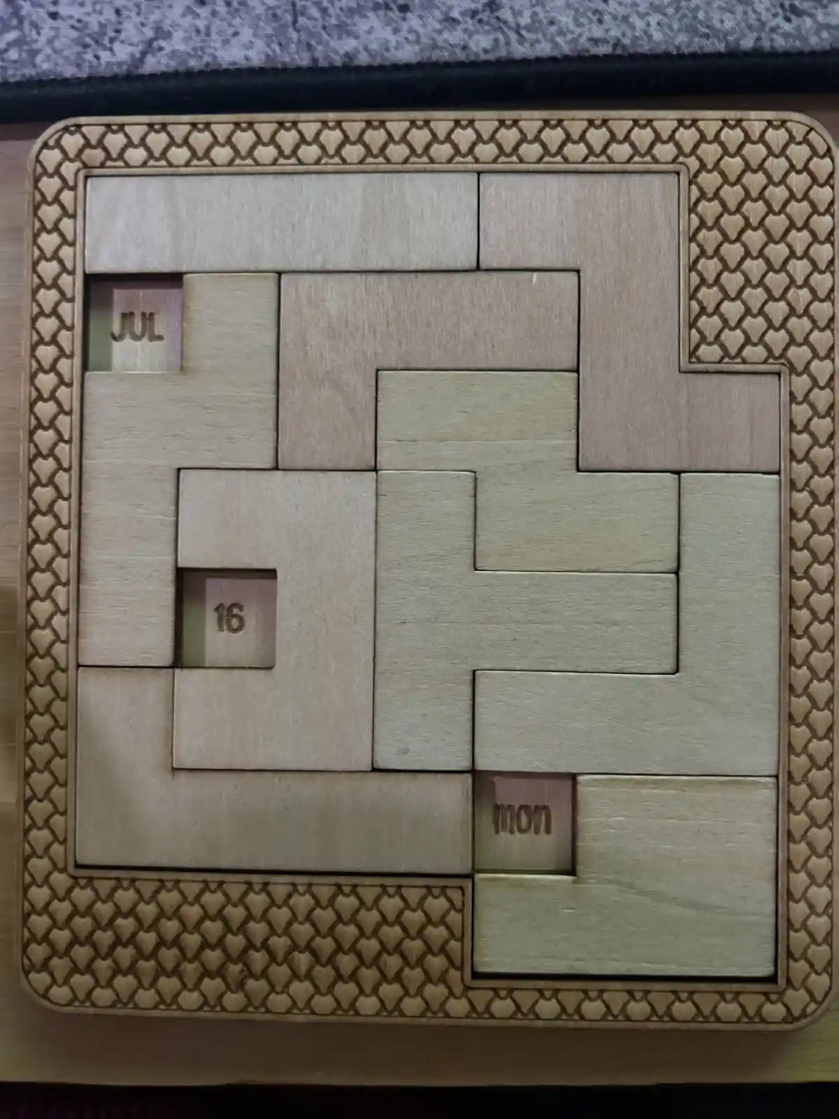 A wooden calendar puzzle with interlocking pieces, showing 'JUL' and '16' in one recessed square and 'MON' in another…