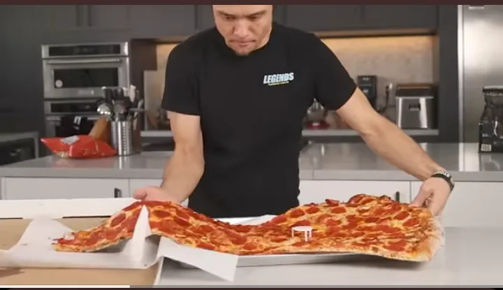 a man in a black t-shirt stands in a modern kitchen holding a very long, thin pepperoni pizza that has been stretched out of…