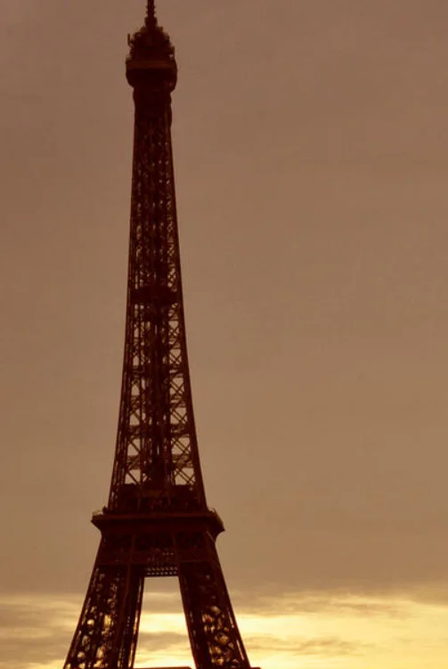 A low-angle shot of the Eiffel Tower silhouetted against a warm, sepia-toned sky at sunset or sunrise. The tower's intricate…