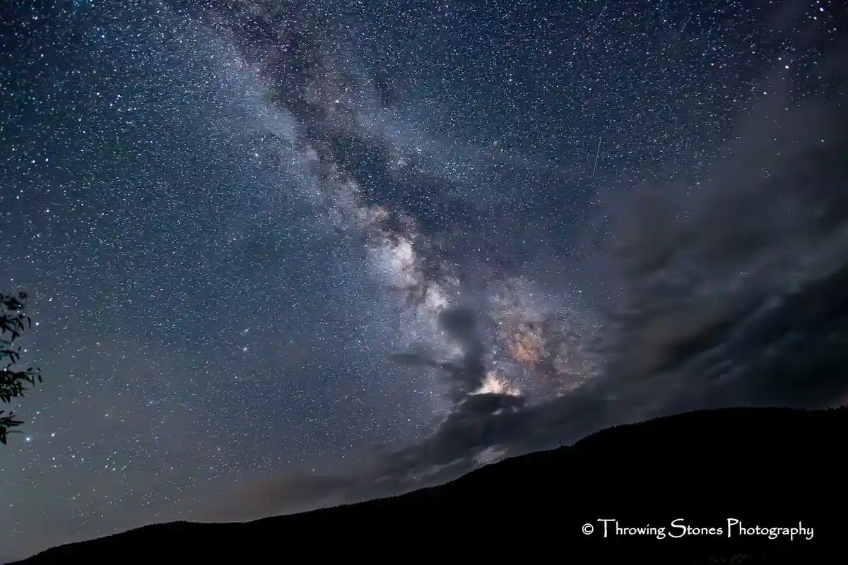 A stunning night sky photograph showing the Milky Way galaxy stretching across a star-filled expanse above dark mountain…