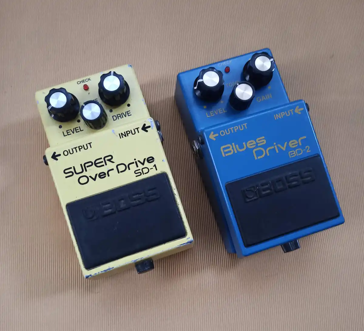 Two Boss guitar pedals, a yellow Super Over Drive SD-1 and a blue Blues Driver BD-2, sit side by side on a brown surface…