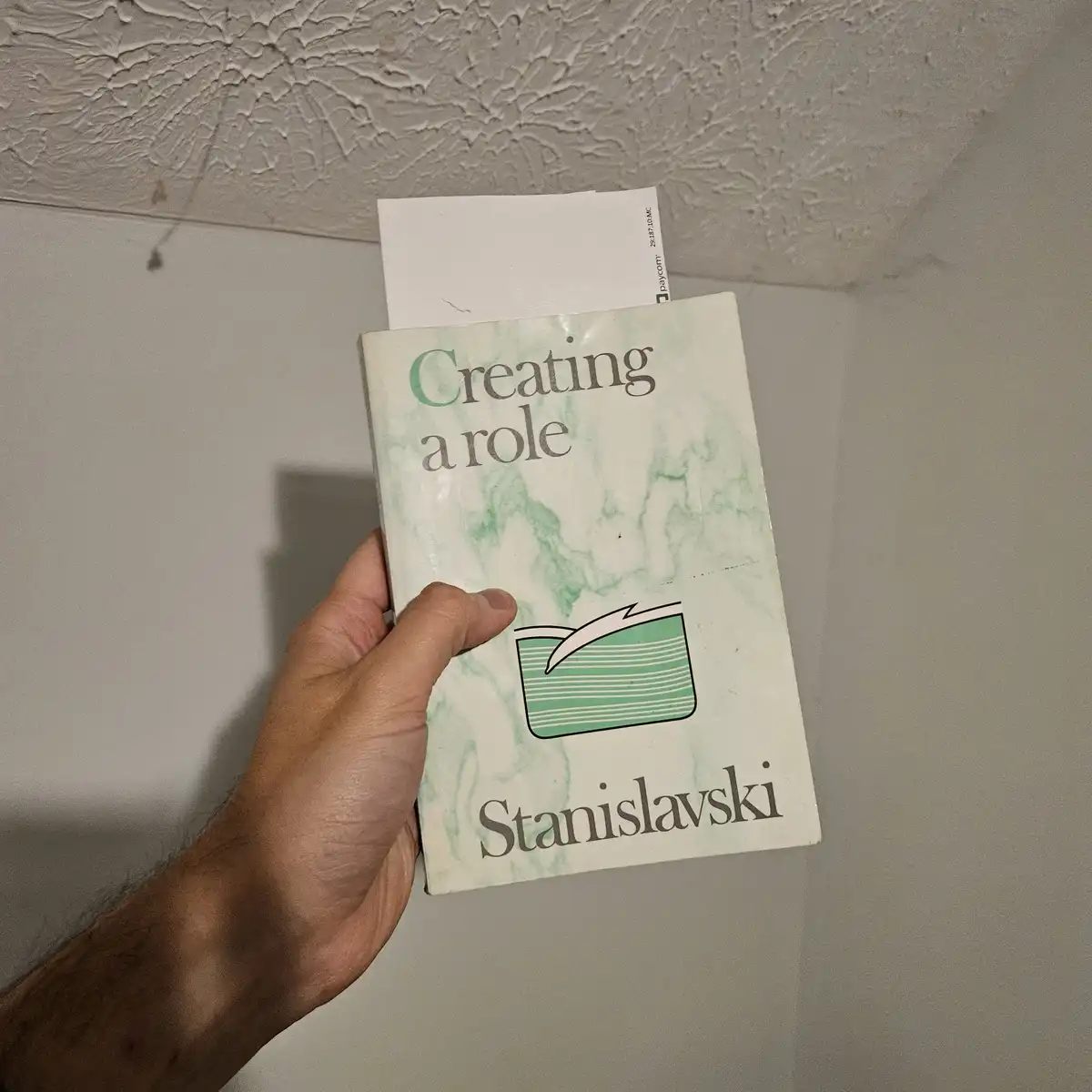 A hand holds a book titled 'Creating a role' by Stanislavski, with a white receipt sticking out from the top. The book cover…