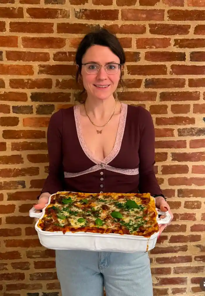A woman with glasses and a maroon top stands in front of a brick wall, holding a white baking dish filled with lasagna…