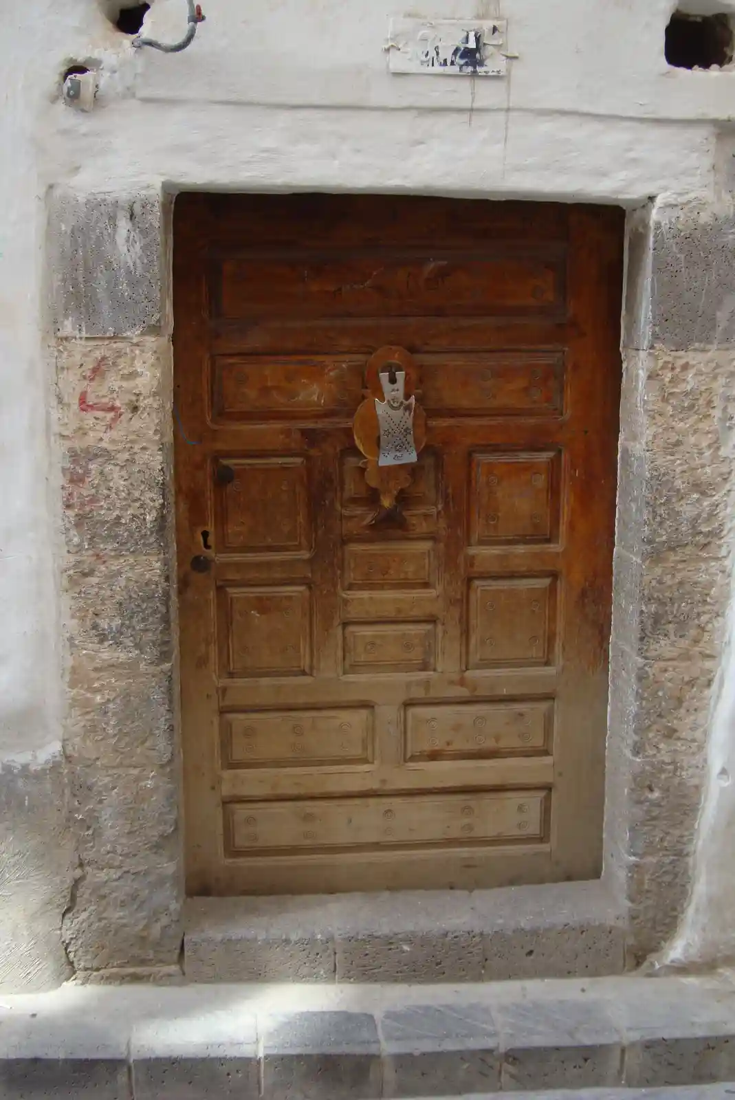 A weathered wooden door with a decorative metal handle is set in a stone and plaster doorway. The door has faint Arabic…