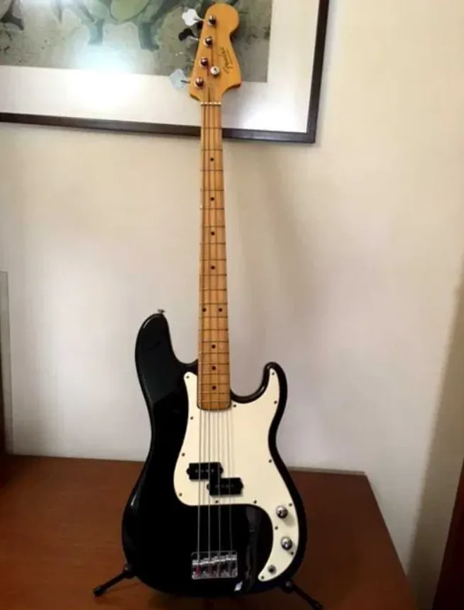A black and white Fender Precision Bass guitar stands upright on a wooden table against a wall with a framed picture above…